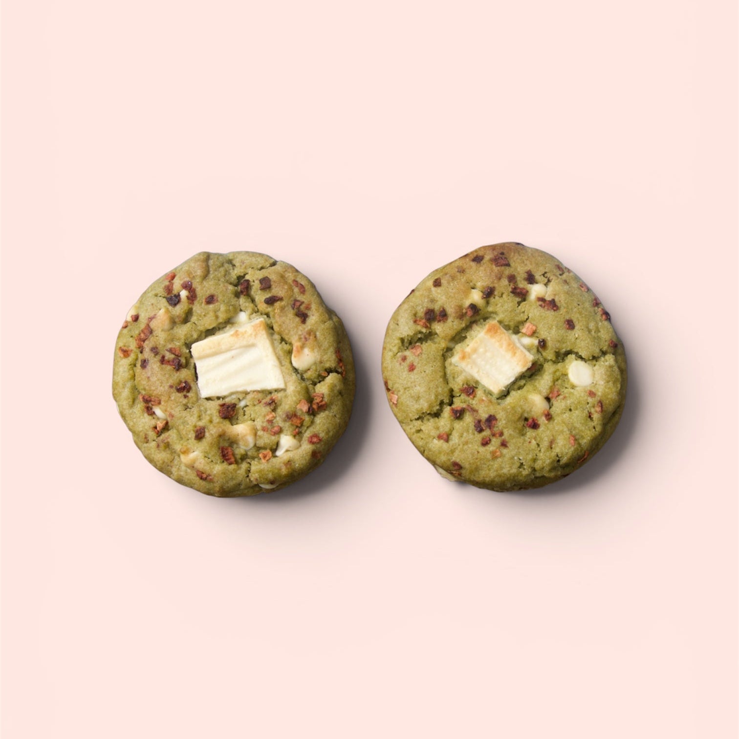 Two green cookies with white chocolate chips on a light pink background